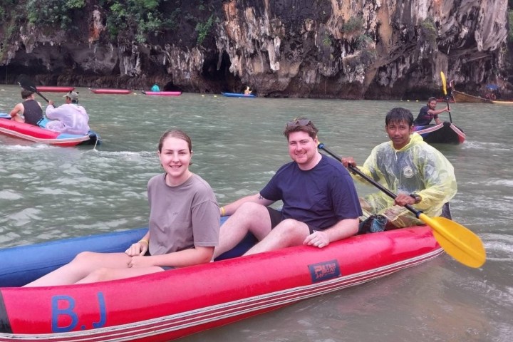 Three people in a red inflatable kayak on a river, with rocky cliffs and greenery in the background.